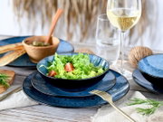 Dunkelblaues Keramik-Geschirrset mit Schüssel und Tellern, darin grüner Salat mit Tomaten, auf gedecktem Holztisch mit Weißweinglas und Besteck, schräg von vorn fotografiert.