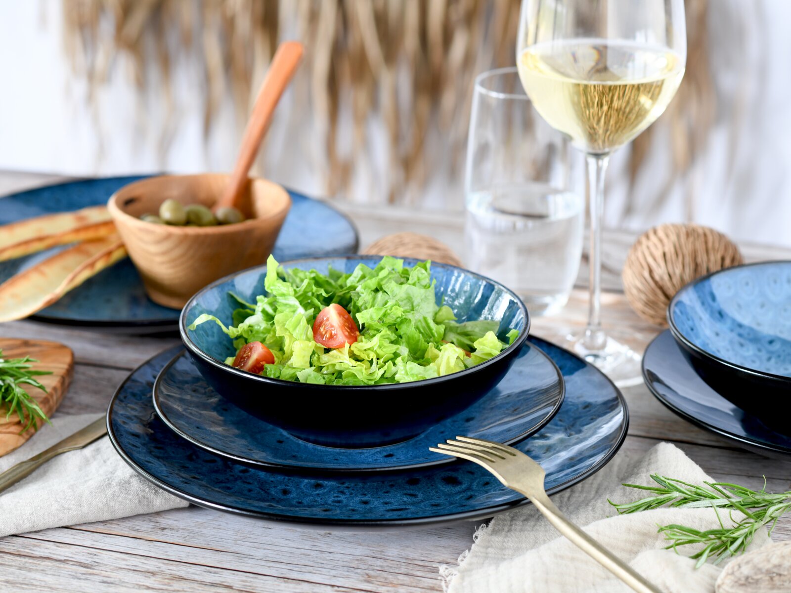 Dunkelblaues Keramik-Geschirrset mit Schüssel und Tellern, darin grüner Salat mit Tomaten, auf gedecktem Holztisch mit Weißweinglas und Besteck, schräg von vorn fotografiert.