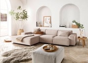 Moderne beige Ecksofa in einem hellen Wohnzimmer mit großen Fenstern, Pflanzen und stilvollen Dekorationen, aus der Perspektive von vorne links.