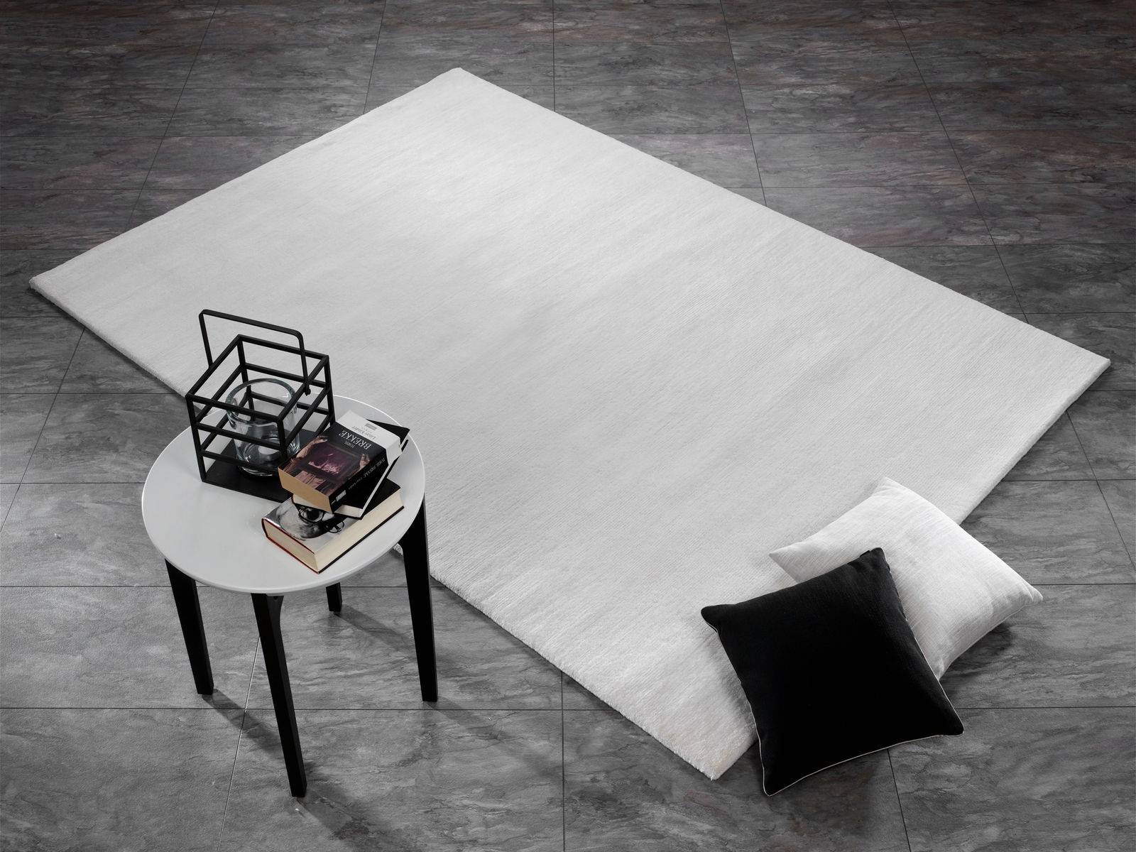 Moderner Wollteppich in hellem Grau auf dunklem Fliesenboden, mit schwarzem und weißem Kissen sowie einem kleinen Tisch mit Büchern und Dekoration. Perspektive von oben.