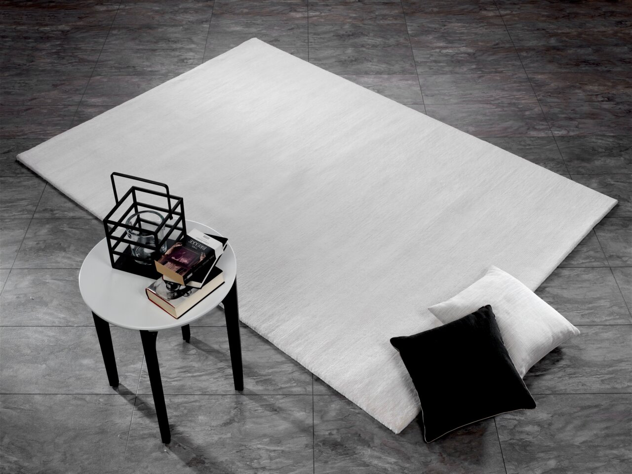 Moderner Wollteppich in hellem Grau auf dunklem Fliesenboden, mit schwarzem und weißem Kissen sowie einem kleinen Tisch mit Büchern und Dekoration. Perspektive von oben.