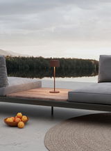 Moderne Akku-Tischleuchte auf einem Holztisch zwischen zwei grauen Sofas mit Blick auf einen ruhigen See und bewaldete Ufer im Hintergrund.