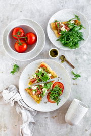 Draufsicht auf einen hellen gedeckten Tisch mit zwei Tellern: belegte Pizzastücke mit Tomaten, Rucola und Käse, dazu frischer Rucola-Salat; daneben eine Schüssel mit Tomaten am Strunk, ein kleines Schälchen Pesto, ein Löffel und ein Becher.
