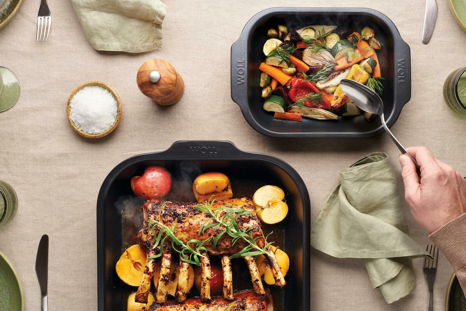 Woll Bratreine ROAST IT Zwei Bratreinen aus schwarzem Material, gefüllt mit gebratenem Gemüse und Fleisch, auf einem gedeckten Tisch von oben betrachtet.
