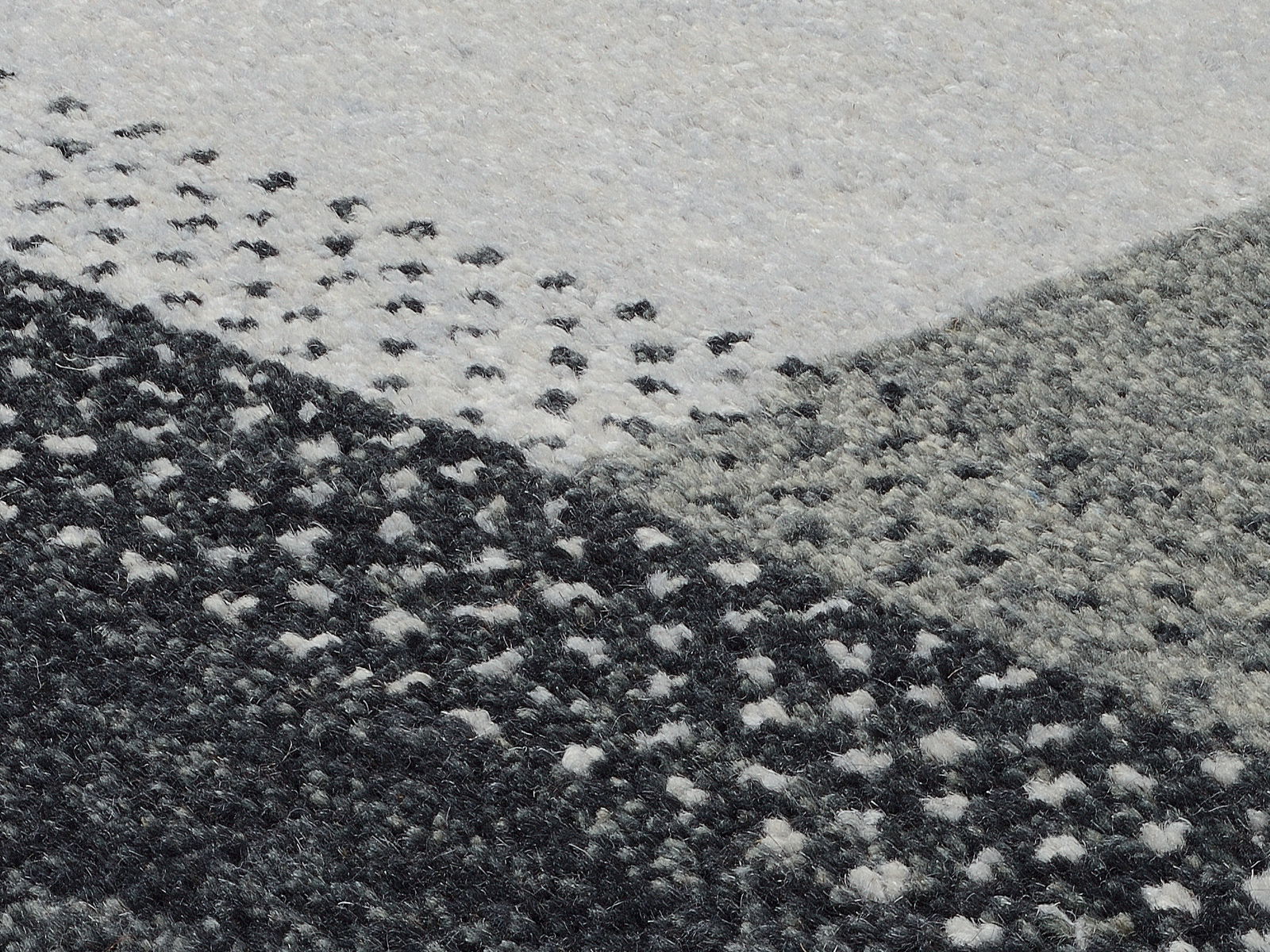 Nahaufnahme eines Wollteppichs mit geometrischem Muster in Grautönen, von oben betrachtet.