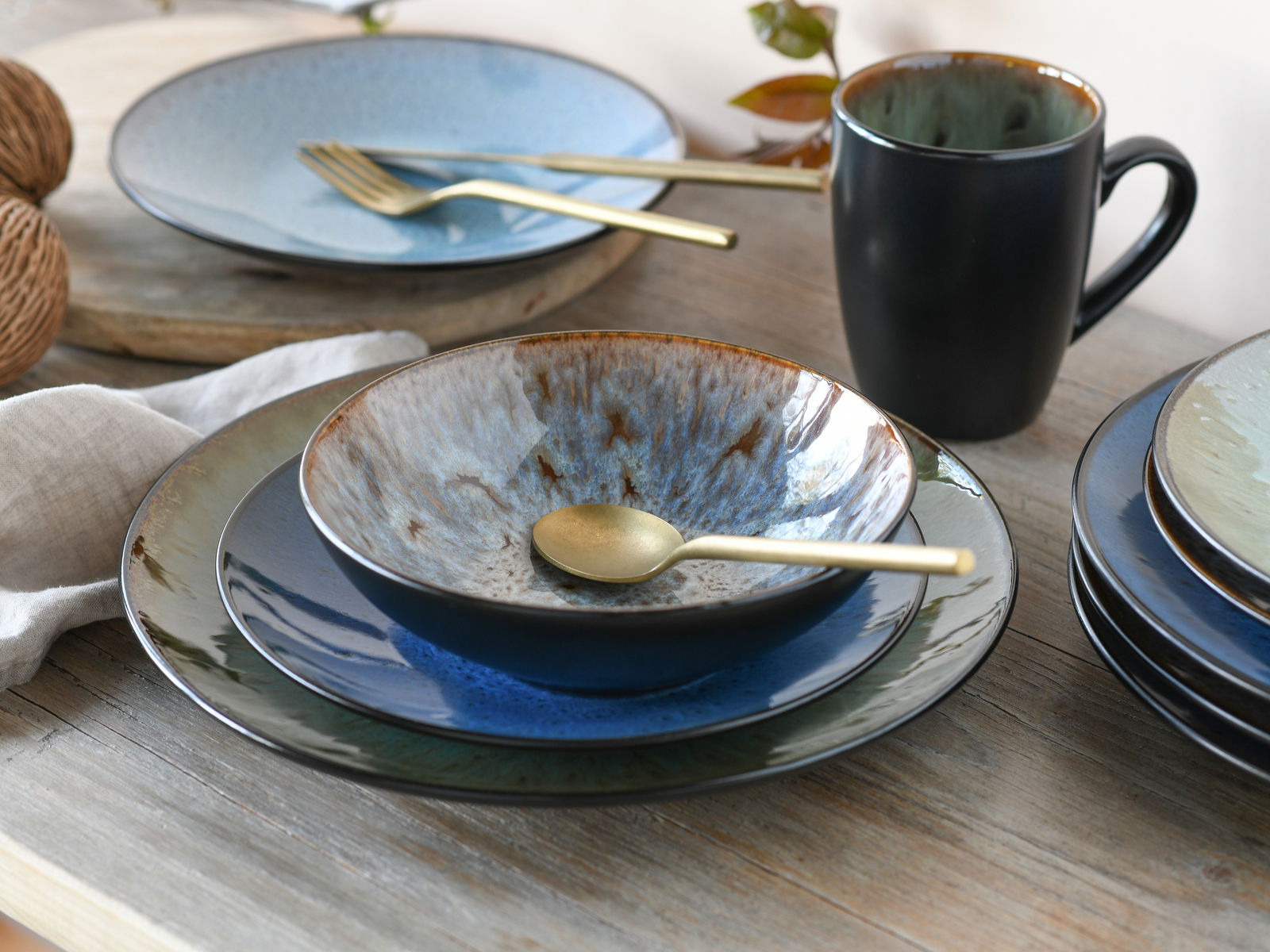 Kombiservice Bali 16-tlg. in Nahaufnahme, bestehend aus Tellern, Schalen und einer Tasse in verschiedenen Blautönen mit goldfarbenem Besteck auf einem Holztisch.