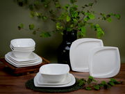 Ein 12-teiliges Tafelservice mit modernen, weißen, quadratischen Tellern und Schalen, präsentiert auf einem Holztisch mit grünen Zweigen im Hintergrund. Die Perspektive zeigt das Set von vorne, wobei die Schalen gestapelt sind.