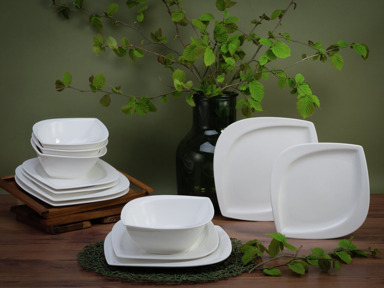 Creatable Tafelservice 12-tlg. MELBOURNE Ein 12-teiliges Tafelservice mit modernen, weißen, quadratischen Tellern und Schalen, präsentiert auf einem Holztisch mit grünen Zweigen im Hintergrund. Die Perspektive zeigt das Set von vorne, wobei die Schalen gestapelt sind.