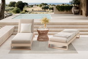 Zwei moderne Outdoor-Liegen in Beige mit Kissen und Decke neben einem runden Beistelltisch auf einer Terrasse am Pool, Blick aus frontaler Perspektive auf die Sitzgruppe mit Landschaft und Meer im Hintergrund