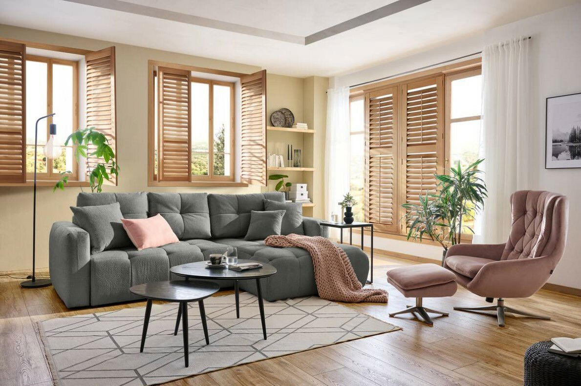 Hom´in Ecksofa London Gemütliche Wohnzimmer-Ecke mit grauem Rundecksofa, dekoriert mit Kissen und Decke, im Vordergrund ein runder Couchtisch, seitliche Perspektive.