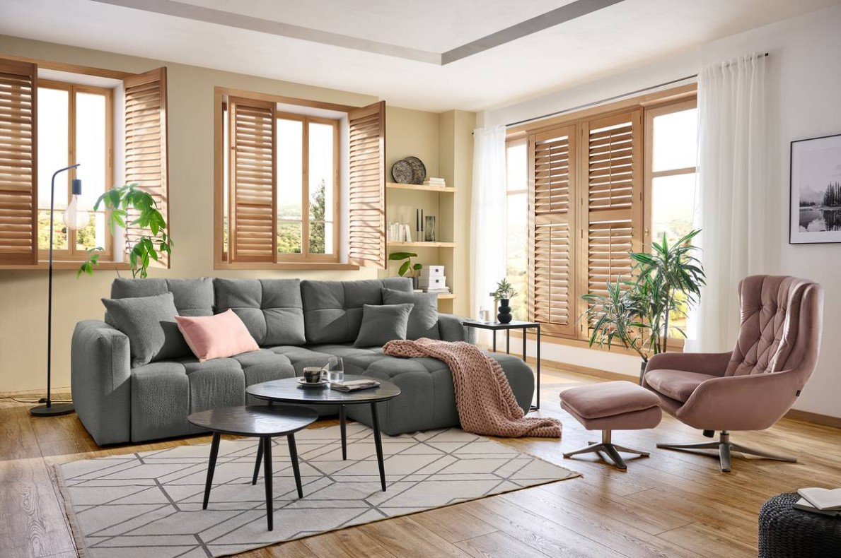 Hom´in Ecksofa London Gemütliche Wohnzimmer-Ecke mit grauem Rundecksofa, dekoriert mit Kissen und Decke, im Vordergrund ein runder Couchtisch, seitliche Perspektive.