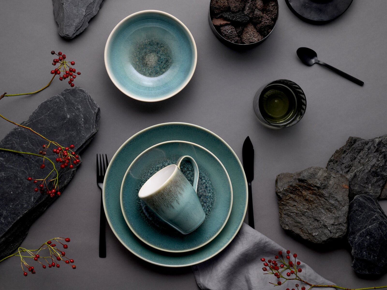 Draufsicht (Flatlay) auf türkisblaues Keramik-Geschirrset mit Schale, Tellern und Tasse, arrangiert auf grauem Hintergrund mit schwarzem Besteck, Steinen und roten Beerenzweigen.