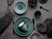 Draufsicht (Flatlay) auf türkisblaues Keramik-Geschirrset mit Schale, Tellern und Tasse, arrangiert auf grauem Hintergrund mit schwarzem Besteck, Steinen und roten Beerenzweigen.