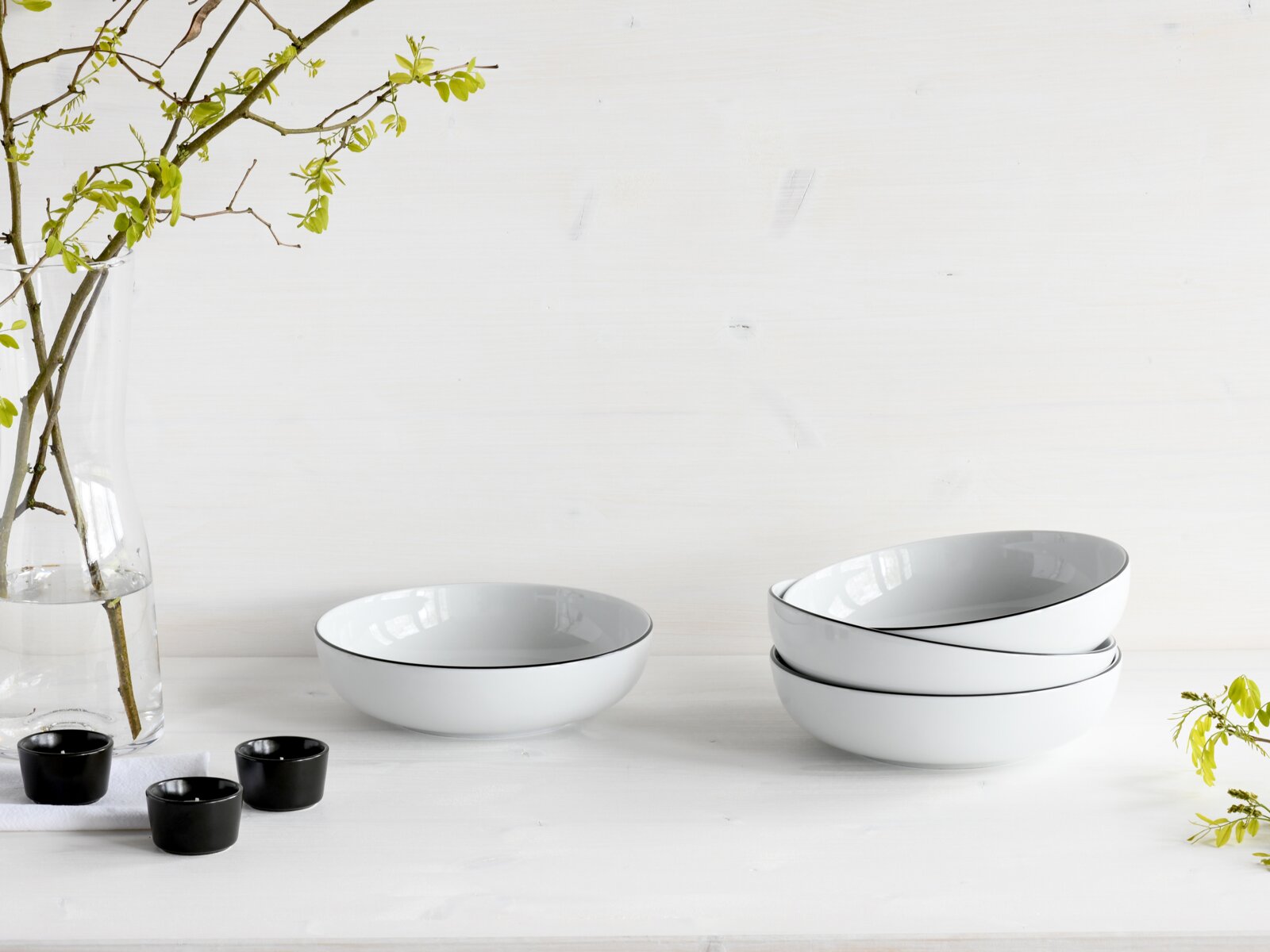 Creatable Poke Bowl 4-tlg ALLROUND Drei weiße, ovale Porzellanschalen mit feiner schwarzer Randlinie auf hellem Tisch, zwei gestapelt und eine einzeln, in leicht schräger Seitenansicht; links Glasvase mit grünen Zweigen und kleine schwarze Schälchen.