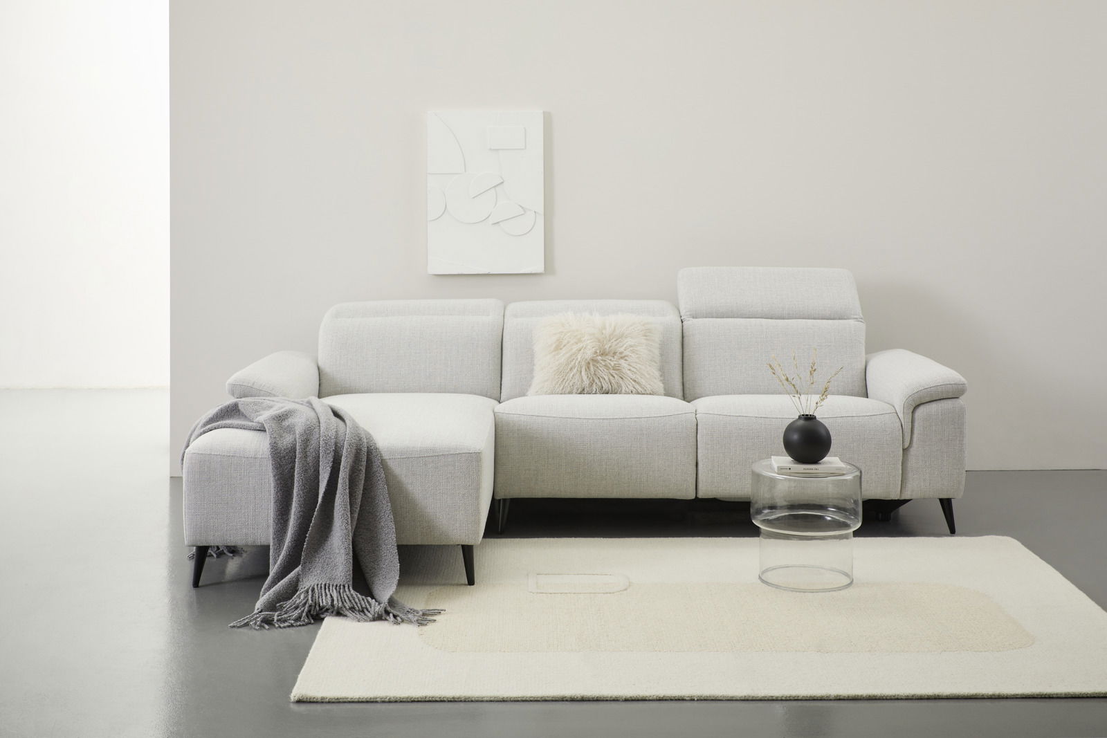 Graue Longchairkombination mit Kissen und Decke, frontal fotografiert, auf hellem Teppich mit rundem Glastisch und schwarzer Vase.