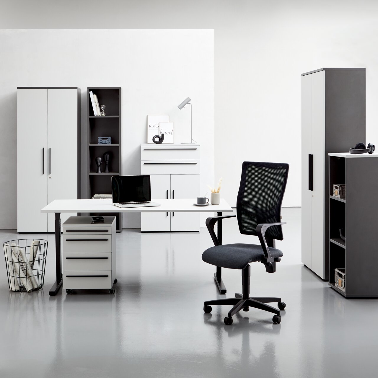 Moderne Büromöbel in einem hellen Raum, darunter ein Schreibtisch mit Rollcontainer, ein Bürostuhl und mehrere Aktenschränke. Frontalansicht.