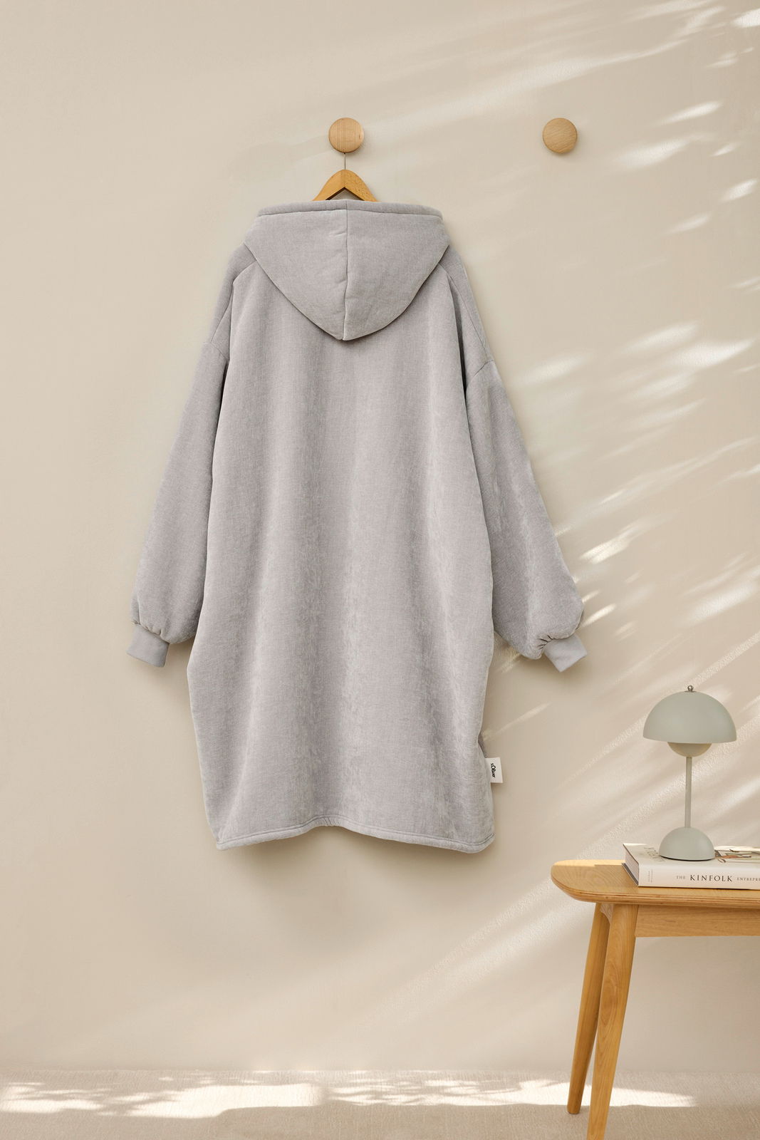 Rückansicht eines grauen Lounge Hoodies, der an einem Holzhaken hängt, mit Kapuze und langen Ärmeln.