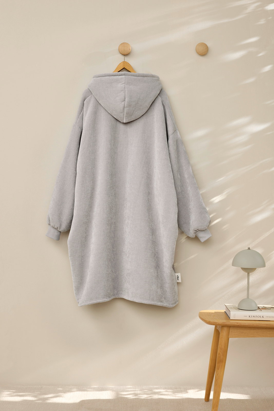 Rückansicht eines grauen Lounge Hoodies, der an einem Holzhaken hängt, mit Kapuze und langen Ärmeln.