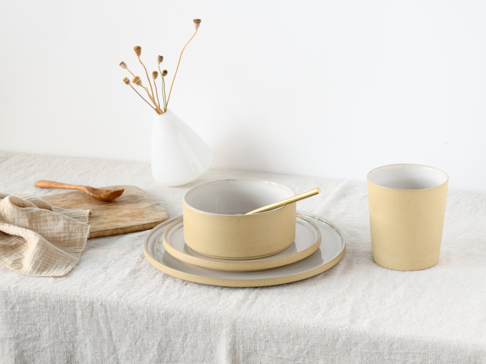 Minimalistisches Keramik-Geschirrset in Creme und Weiß mit Schale auf Tellern und Becher auf Leinentuch, schräg von vorne fotografiert.