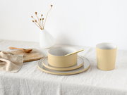 Minimalistisches Keramik-Geschirrset in Creme und Weiß mit Schale auf Tellern und Becher auf Leinentuch, schräg von vorne fotografiert.