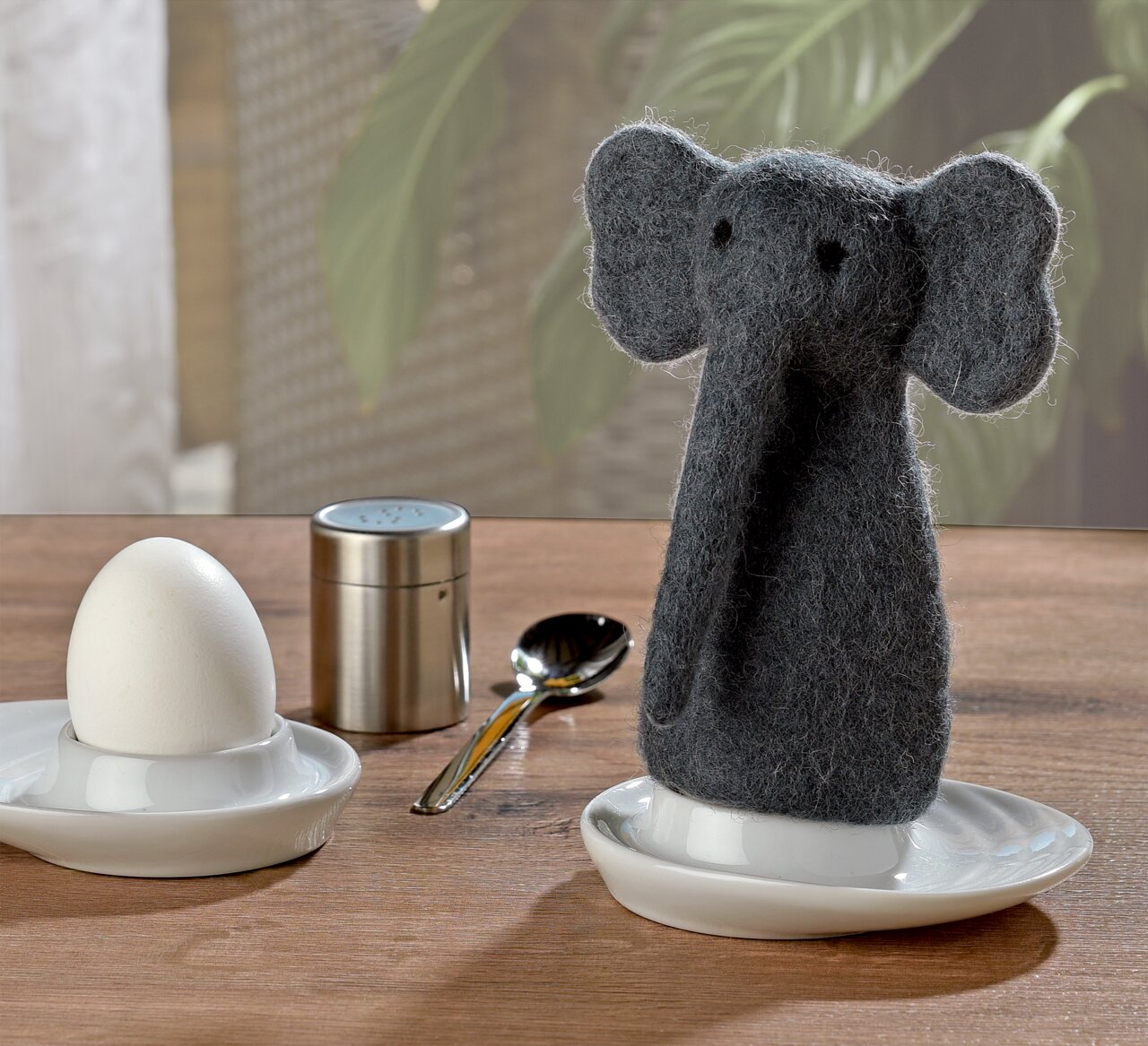 Eierwärmer in Form eines grauen Elefanten aus Filz, frontal auf einem Tisch neben einem Ei auf einem Eierbecher, mit einem Löffel und einem Salzstreuer im Hintergrund.