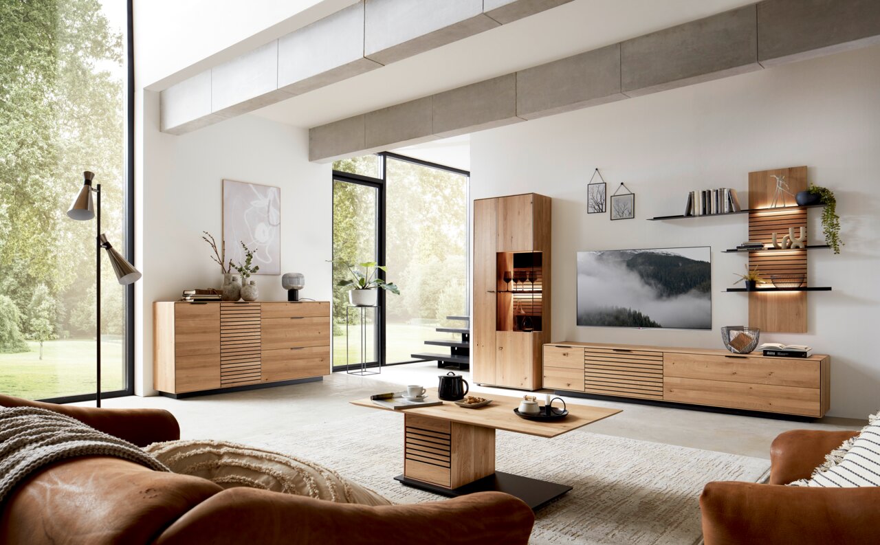 Modernes Wohnzimmer mit einem Sideboard aus Holz, das von der Seite betrachtet wird. Das Sideboard hat eine glatte Oberfläche und bietet Stauraum mit Schubladen und Türen. Es steht in einem hellen Raum mit großen Fenstern und stilvoller Dekoration.