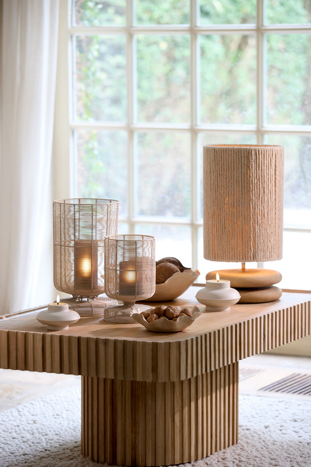 Ein stilvoll dekorierter Tisch mit einer Schale voller Nüsse im Vordergrund, umgeben von Kerzen und einer Lampe, mit einem großen Fenster im Hintergrund. Perspektive von schräg oben.