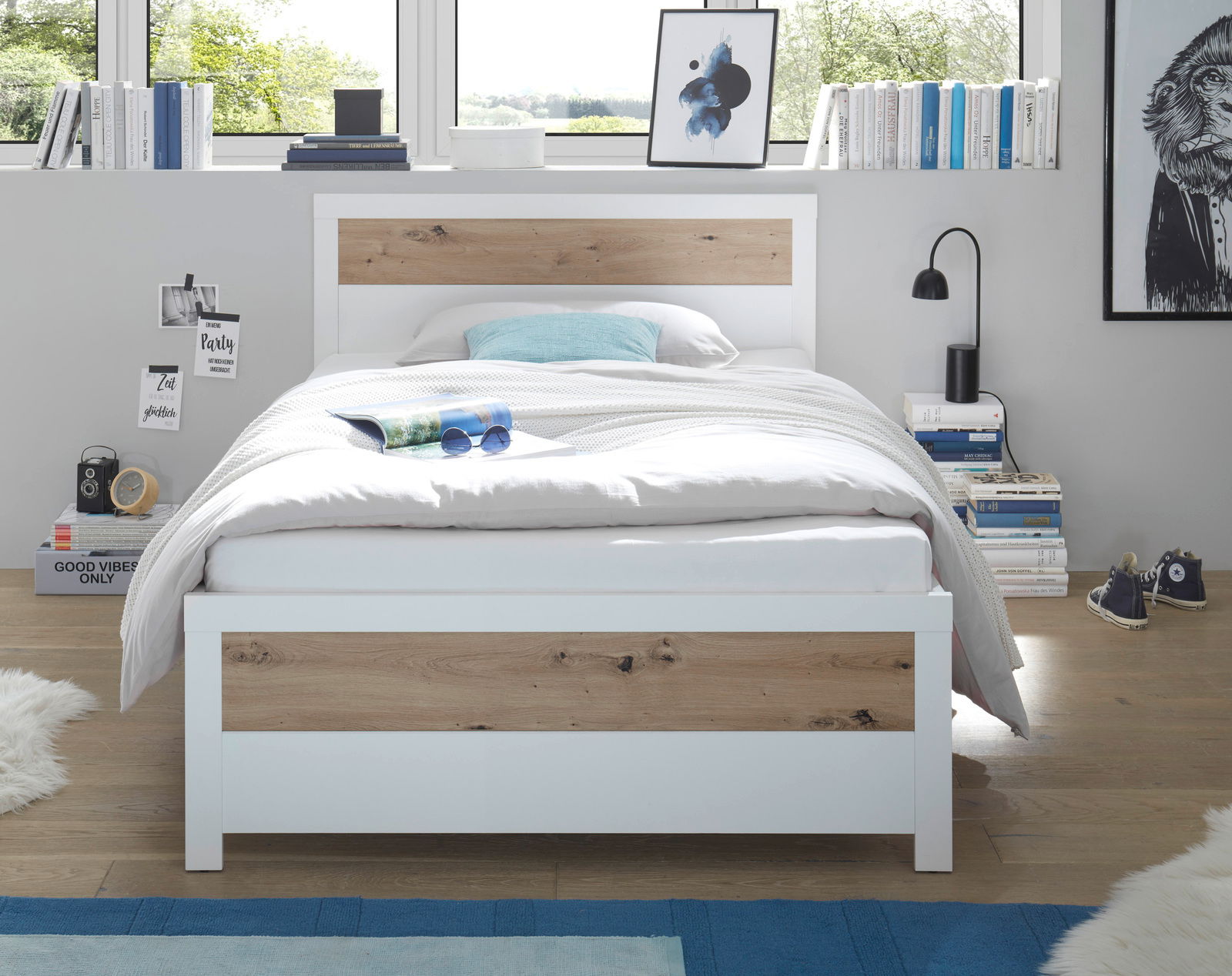 Modernes Bett mit weißem Rahmen und Holzakzenten, frontal fotografiert in einem hellen Raum mit großen Fenstern.