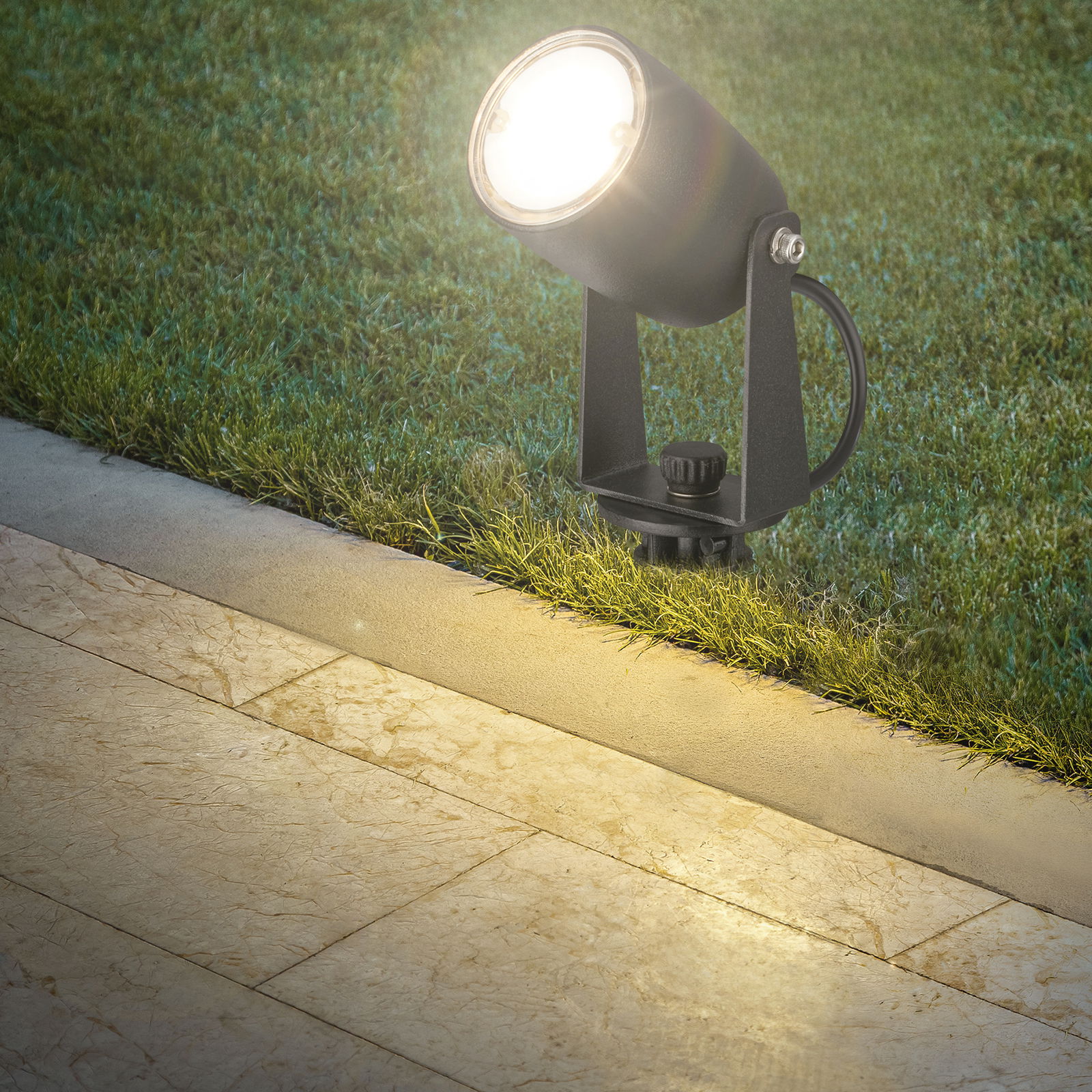 Erdspießstrahler leuchtet von der Seite auf eine Terrasse und Rasenfläche.