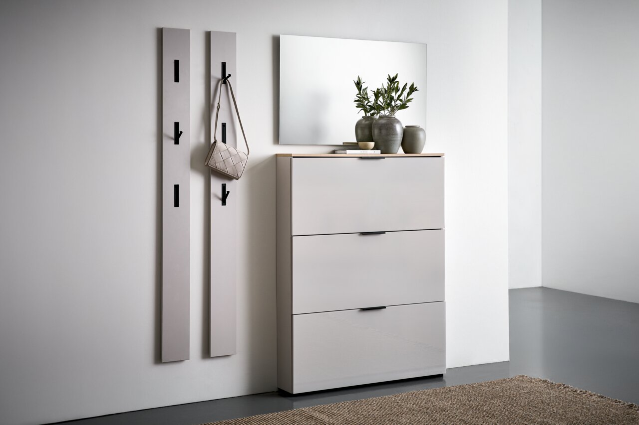 Moderne Garderobeneinrichtung mit zwei vertikalen Paneelen mit Haken, einem Spiegel und einem dreitürigen Schuhschrank in einer hellen, minimalistischen Umgebung. Perspektive von vorne rechts.