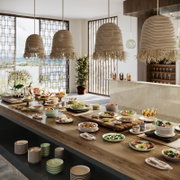 Moderne Buffet-Szene mit reich gedecktem Tisch und mehreren rattanartigen Pendelleuchten mit Fransen in einem hellen Raum, seitliche Perspektive schräg von vorn auf den Tisch