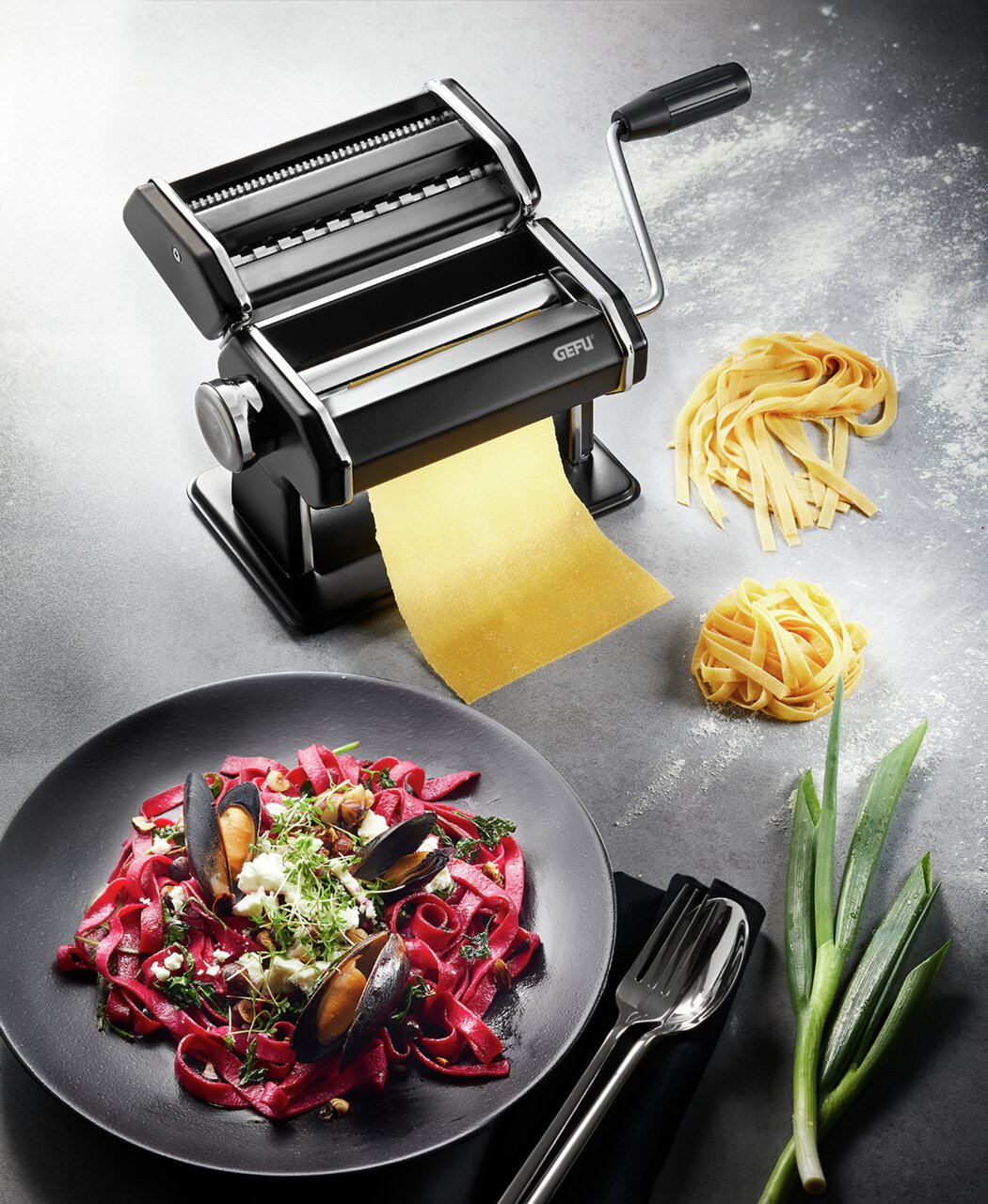 Zwarte pastamachine in mat met slinger, die verse pasta maakt, gefotografeerd vanuit vogelperspectief. Ernaast een bord met rode pasta en schelpdieren, evenals verse ingrediënten.
