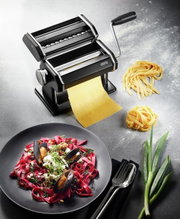 Zwarte pastamachine in mat met slinger, die verse pasta maakt, gefotografeerd vanuit vogelperspectief. Ernaast een bord met rode pasta en schelpdieren, evenals verse ingrediënten.