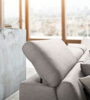 Nahaufnahme einer modernen, hellgrauen Rundecke mit verstellbarer Rückenlehne in einem hellen Raum mit großen Fenstern.