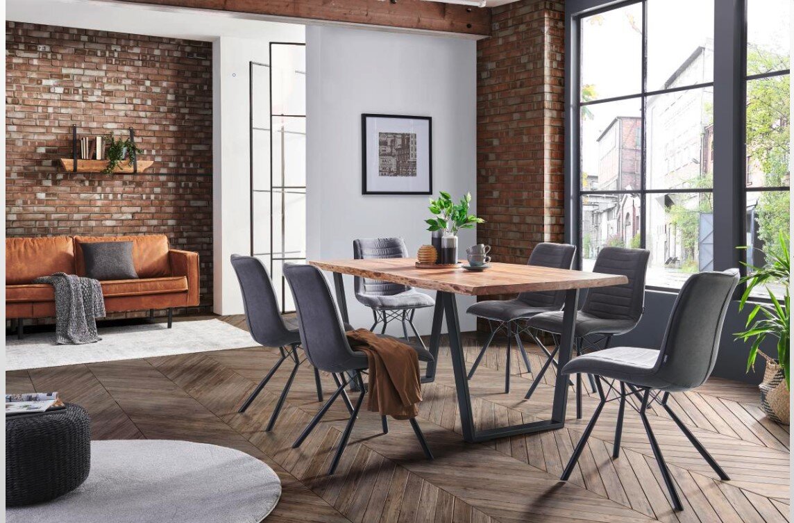 Eetkamer met een moderne tafel met V-vormig onderstel, omringd door grijze stoelen, in een ruimte met een bakstenen muur en grote ramen. Aanzicht vanaf de zijkant.
