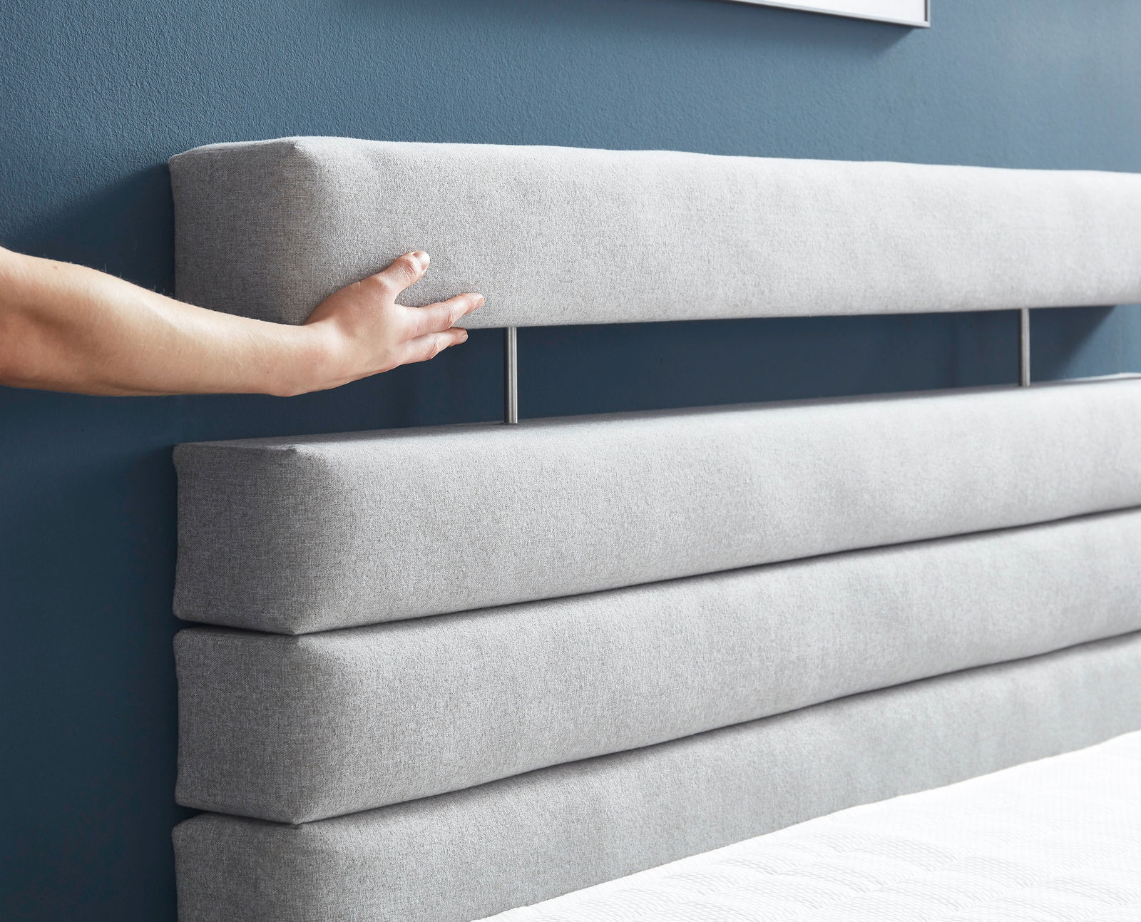 Nahaufnahme eines grauen Boxspringbett-Kopfteil mit horizontalen Polsterstreifen, Hand berührt das Kopfteil.