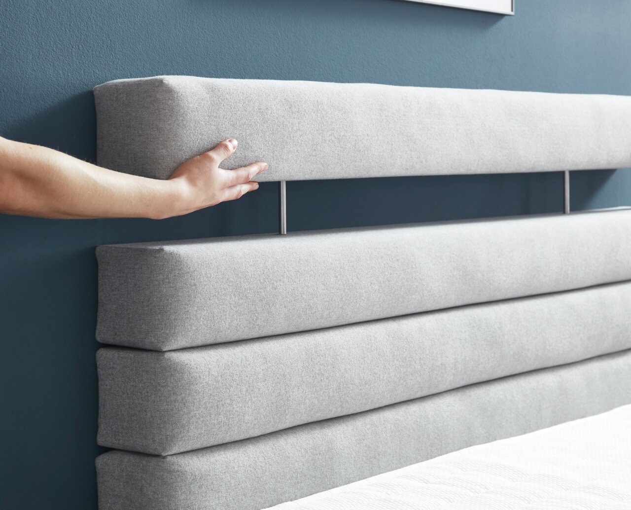 Nahaufnahme eines grauen Boxspringbett-Kopfteil mit horizontalen Polsterstreifen, Hand berührt das Kopfteil.
