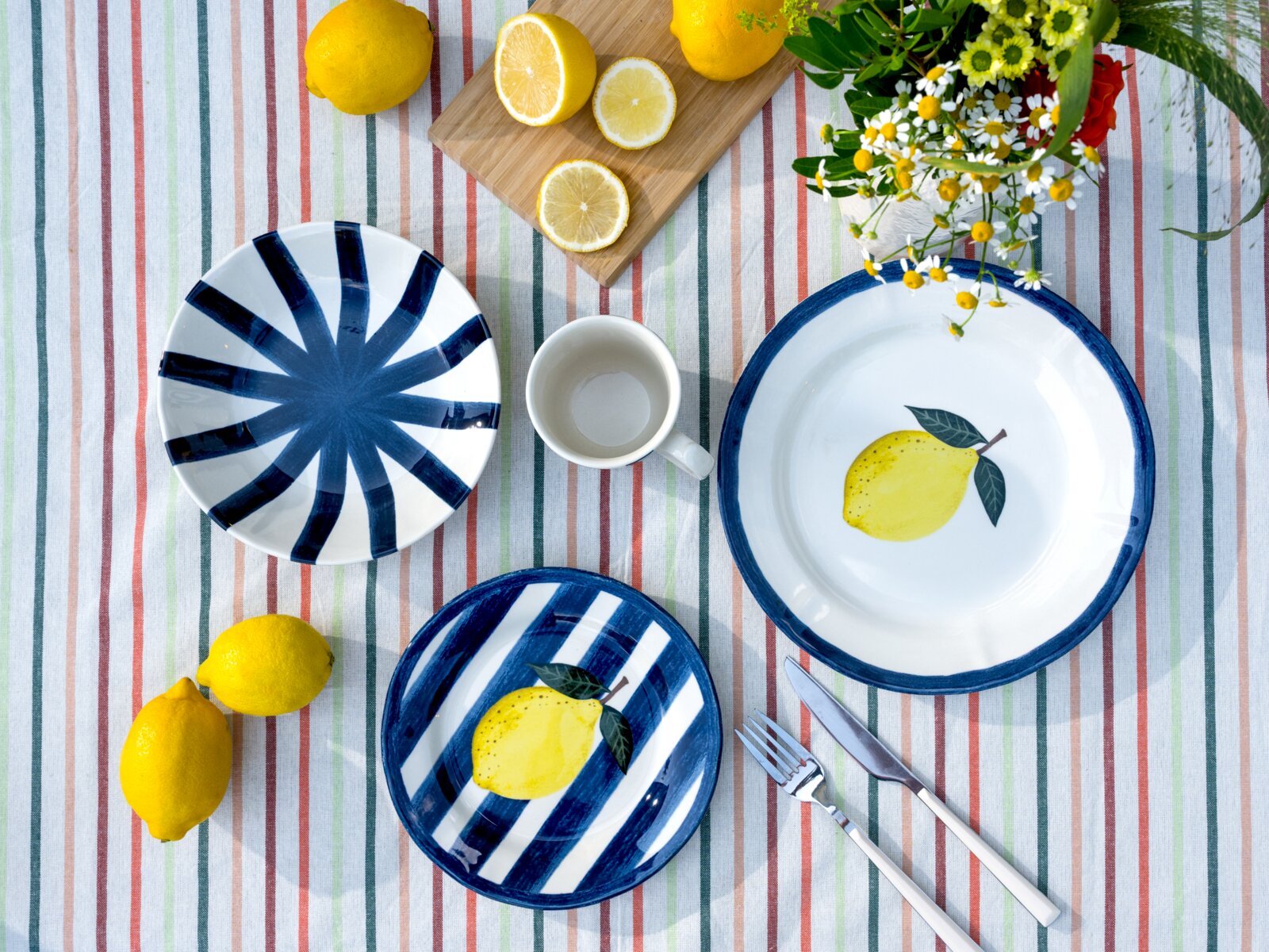 Draufsicht auf blau-weißes Geschirrset mit Zitronenmotiv (Teller und Schale) sowie gestreiftem Teller und weißer Tasse auf bunt gestreifter Tischdecke, dekoriert mit frischen Zitronen, Besteck und Blumen.