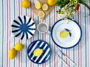 Draufsicht auf blau-weißes Geschirrset mit Zitronenmotiv (Teller und Schale) sowie gestreiftem Teller und weißer Tasse auf bunt gestreifter Tischdecke, dekoriert mit frischen Zitronen, Besteck und Blumen.