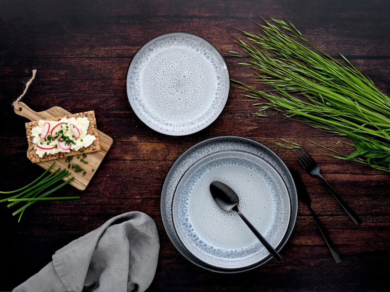 Draufsicht auf ein 18-teiliges Tellerset 'Frost' mit elegantem, gesprenkeltem Design in Grau- und Blautönen, arrangiert mit Besteck, einem Holzbrett mit belegtem Knäckebrot und frischen Kräutern auf einem dunklen Holztisch.