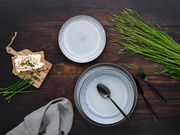 Draufsicht auf ein 18-teiliges Tellerset 'Frost' mit elegantem, gesprenkeltem Design in Grau- und Blautönen, arrangiert mit Besteck, einem Holzbrett mit belegtem Knäckebrot und frischen Kräutern auf einem dunklen Holztisch.