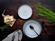 Draufsicht auf ein 18-teiliges Tellerset 'Frost' mit elegantem, gesprenkeltem Design in Grau- und Blautönen, arrangiert mit Besteck, einem Holzbrett mit belegtem Knäckebrot und frischen Kräutern auf einem dunklen Holztisch.