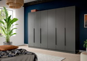 Großer moderner Kleiderschrank in mattem Grau mit sechs Türen und langen schwarzen Griffen, in einem Schlafzimmer vor dunkelblauer Wand, perspektivisch in leichter Schrägansicht von vorn.
