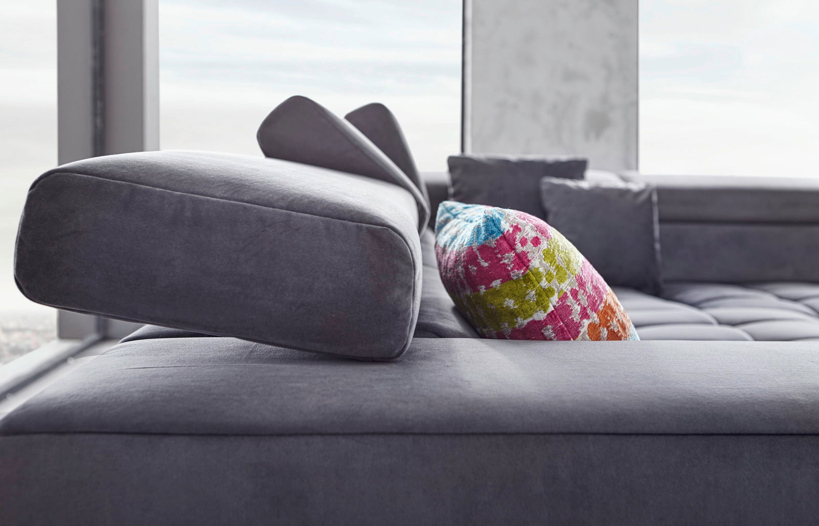 Nahaufnahme einer modernen Polsterrundecke in Grau mit verstellbarer Kopfstütze und buntem Kissen, seitliche Perspektive.