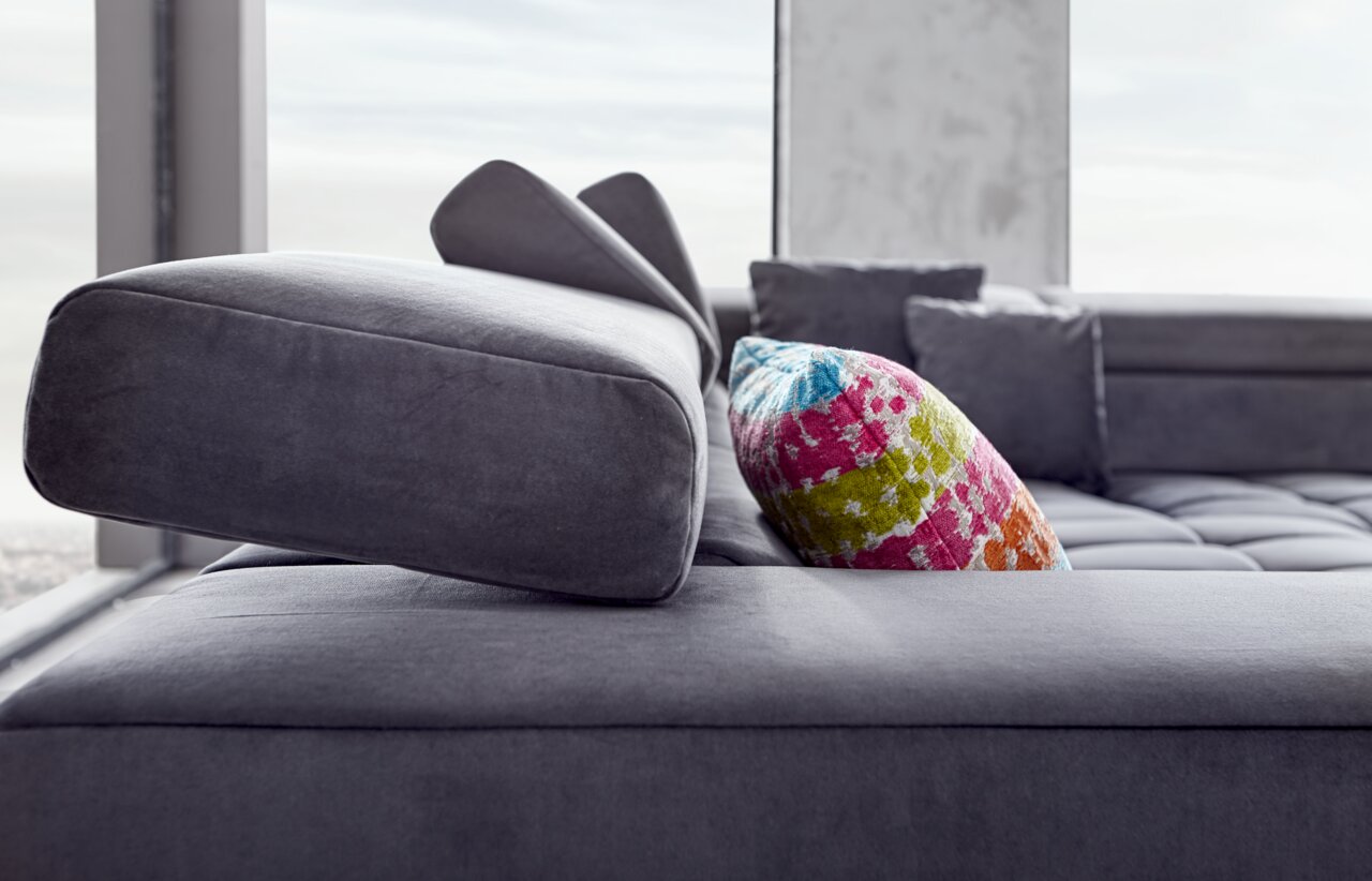 Nahaufnahme einer modernen Polsterrundecke in Grau mit verstellbarer Kopfstütze und buntem Kissen, seitliche Perspektive.