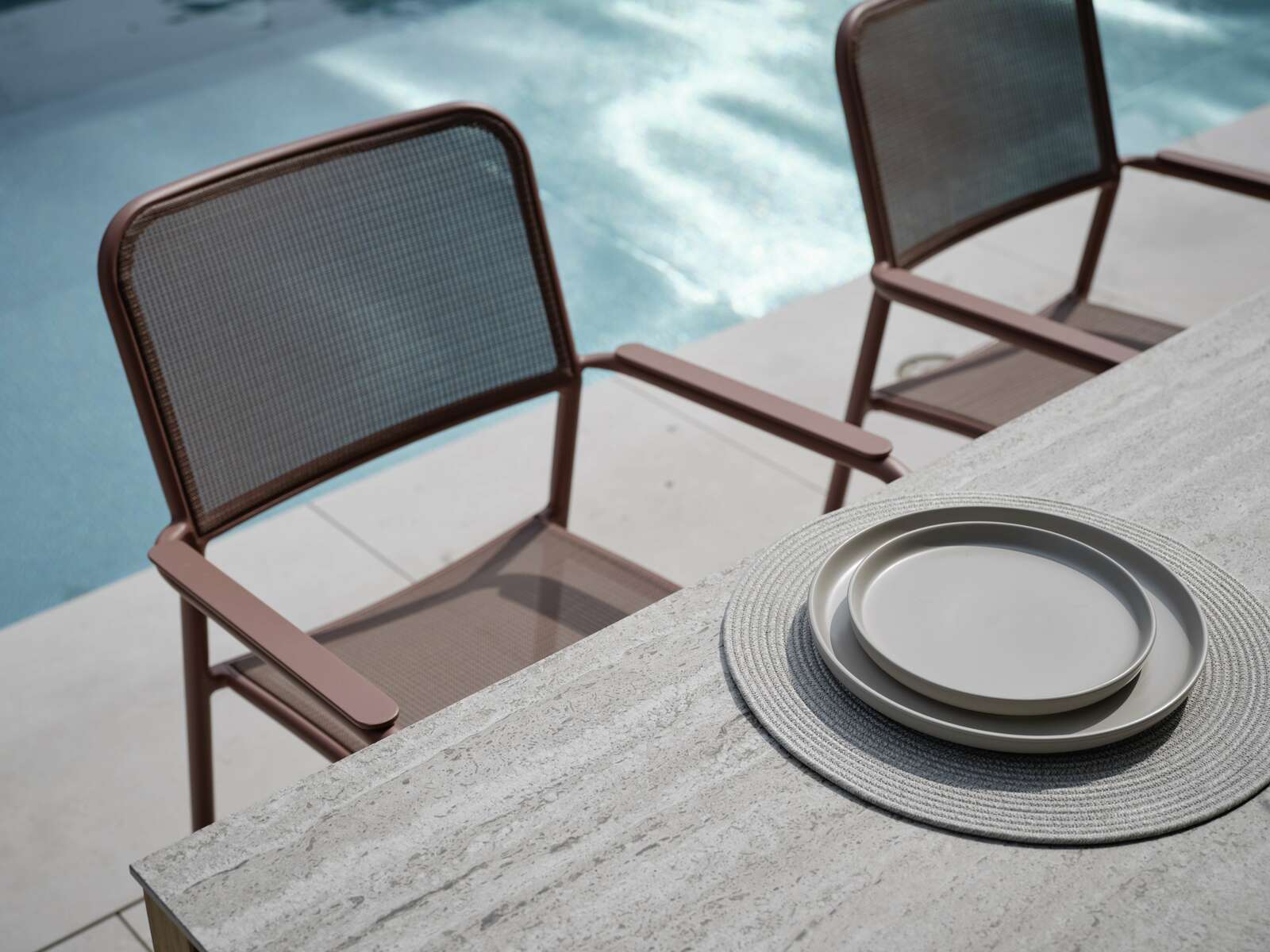 Zwei braune Stapelsessel aus Metall mit Netzgewebe, aufgestellt neben einem Pool, mit Blick von oben auf einen Tisch mit grauen Tellern.