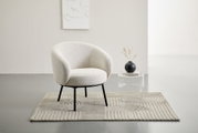 Heller, gepolsterter Loungesessel in Creme/Beige mit schwarzem Metallgestell auf strukturiertem Teppich, frontale Perspektive im minimalistischen Wohnraum neben transparentem Beistelltisch mit schwarzer Vase.