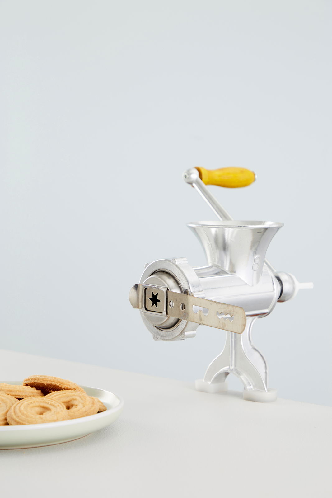 Fleischwolf Nr. 8 aus Metall mit gelbem Holzgriff, seitliche Perspektive, neben einem Teller mit Keksen.