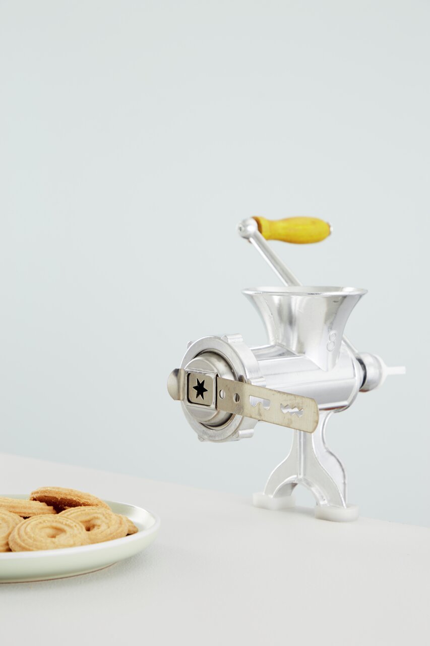 Fleischwolf Nr. 8 aus Metall mit gelbem Holzgriff, seitliche Perspektive, neben einem Teller mit Keksen.