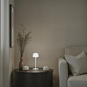 Weiße, minimalistische Tischleuchte mit rundem Schirm auf schwarzem, rundem Beistelltisch neben grauem Sofa; Frontalansicht auf Augenhöhe.